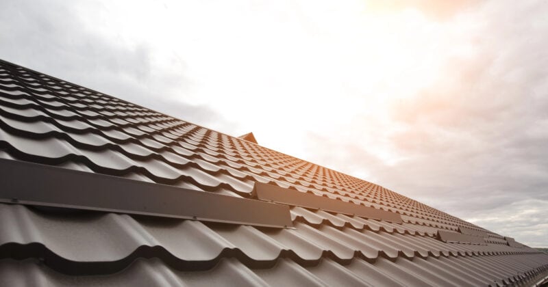 A sunlit metal roof with a wavy sheet pattern extends toward a cloudy sky, reflecting sunlight near the top—an ideal showcase for effective roofing marketing, drawing eyes and attracting more clients with its captivating design.