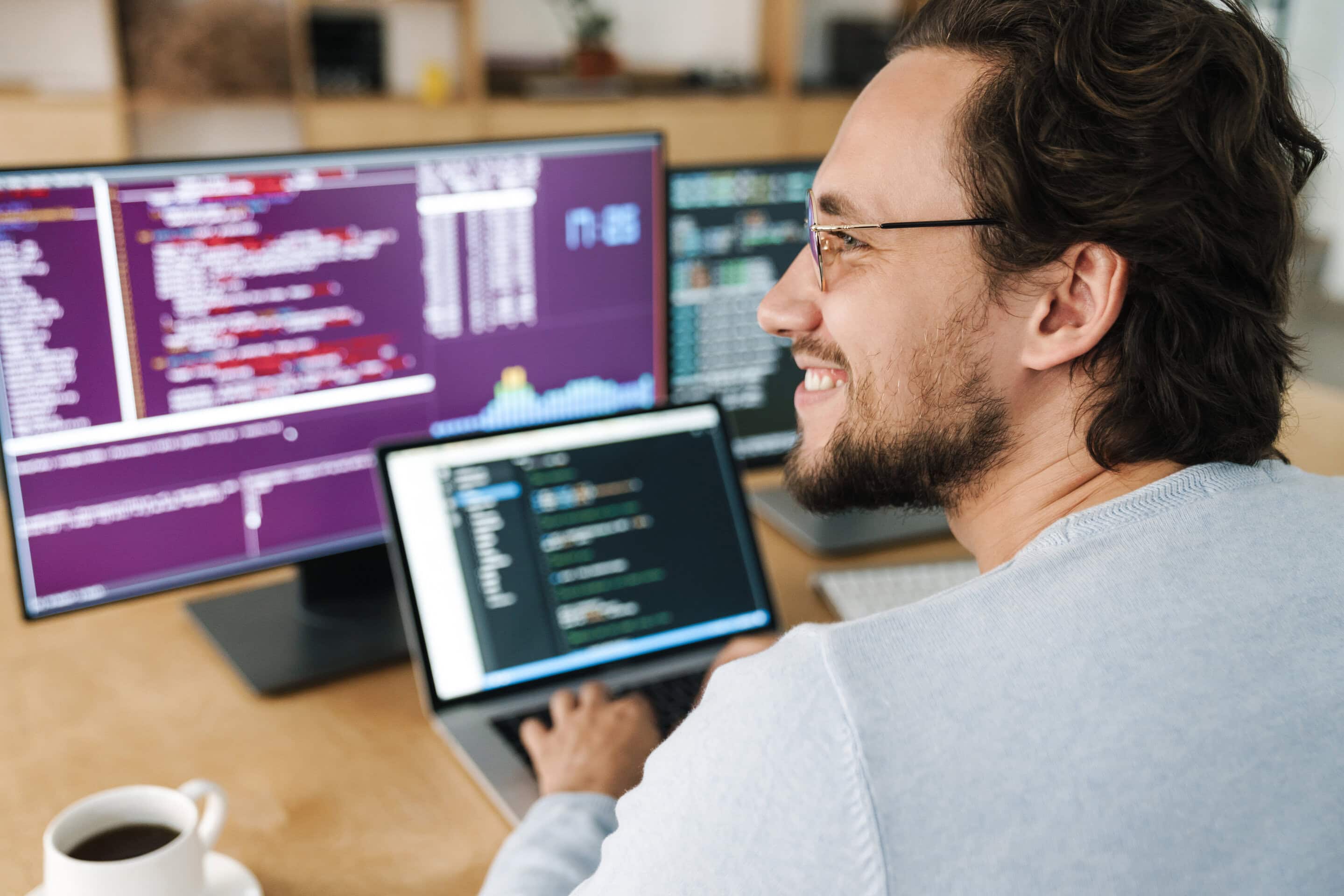 A smiling person wearing glasses works at a desk with two monitors and a laptop displaying code for HTML to WordPress site conversion. A cup of coffee sits nearby, and the background shows shelves with books and decor.
