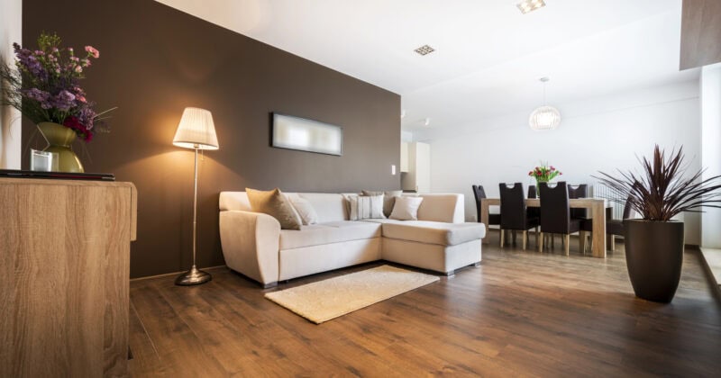 Discover modern living room inspiration for 2025 with wooden flooring, a beige sectional sofa, and a floor lamp. A dining table set graces the background, while a brown accent wall and potted plants provide decorative touches. Learn from the best on leading home decor websites.
