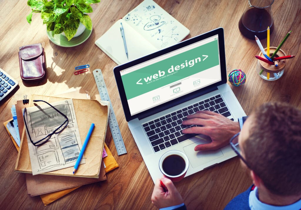 The person is focused on web design using a laptop, with coffee, design sketches, and papers scattered around. A plant adds a touch of nature to the wooden desk as they manage content for a seamless WordPress Migration.