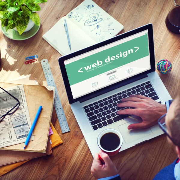 The person is focused on web design using a laptop, with coffee, design sketches, and papers scattered around. A plant adds a touch of nature to the wooden desk as they manage content for a seamless WordPress Migration.