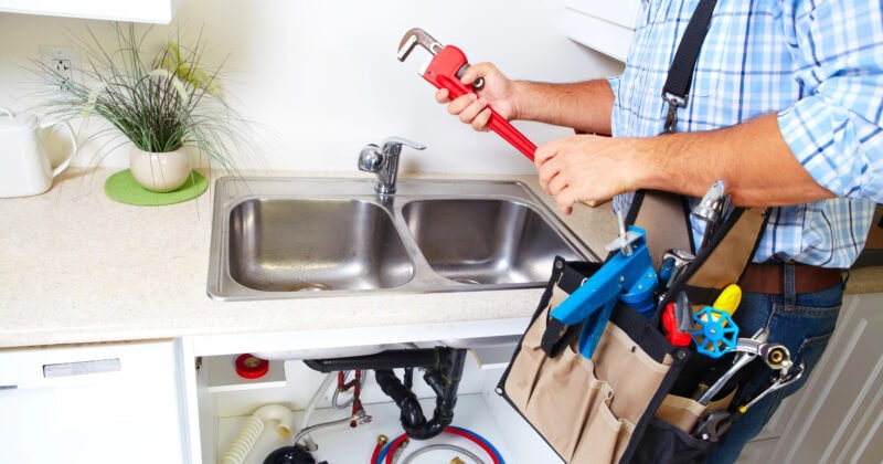 Person holding a wrench stands by a kitchen sink with a toolbag, engaging in repairs. The cabinet below the sink is open, revealing plumbing tools and pipes—an ideal scene for showcasing how effective plumbing website design converts visitors into clients.