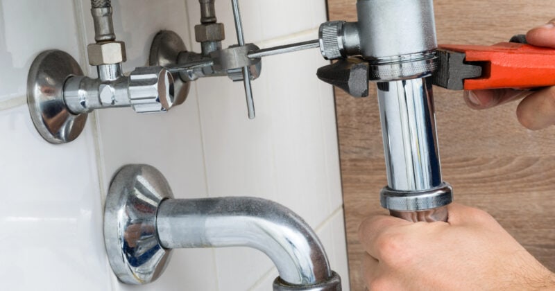 In a competitive market, a person deftly uses a wrench to tighten a pipe under the sink, surrounded by an array of plumbing fixtures.