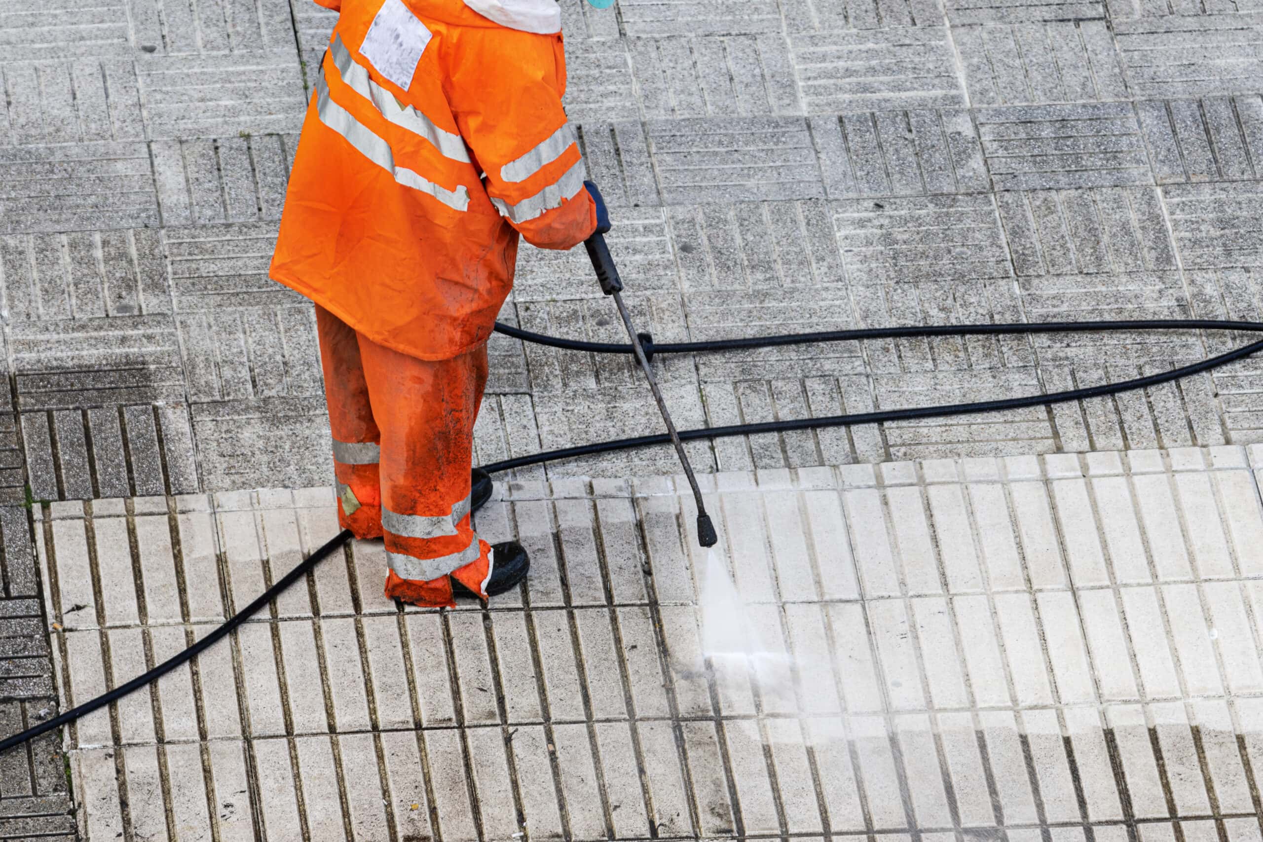 A person wearing an orange reflective suit uses a pressure washer to clean patterned pavement tiles, revealing a lighter, cleaner surface—like how A/B Testing in Plumbing Facebook Ads helps maximize budget and achieve standout results.