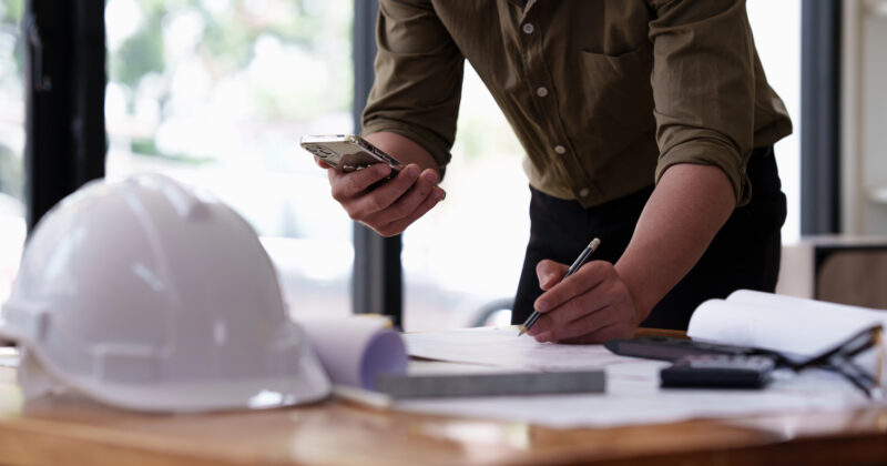A person in a green shirt is using a smartphone to manage Contractor Ads while jotting down notes at a desk, where a white hard hat sits nearby.
