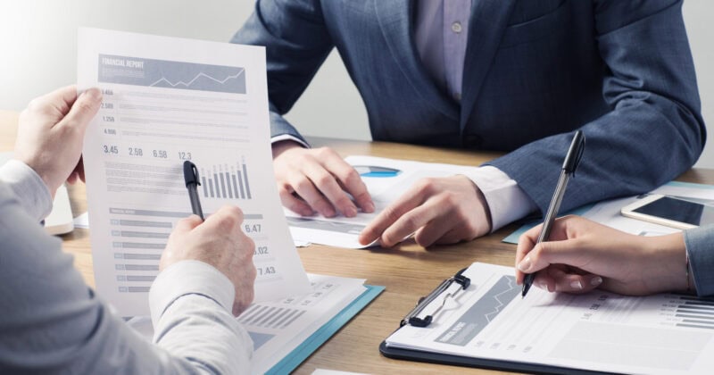 Three financial advisors in business attire sit at a table, reviewing documents and reports. One holds a pen, gesturing at a document while the others take notes, building trust through detailed analysis.