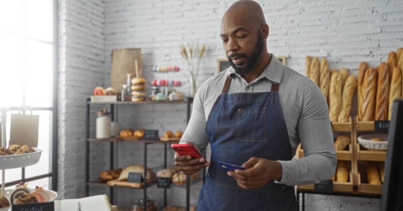 A man in an apron stands in a bakery, looking at his phone while holding a credit card. Racks of bread and pastries are displayed in the background.