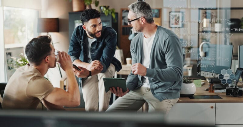 Three men discussing around a table in an office, with one writing on a glass board, brainstorming marketing strategies. They appear engaged in conversation, and various office supplies are visible in the background.