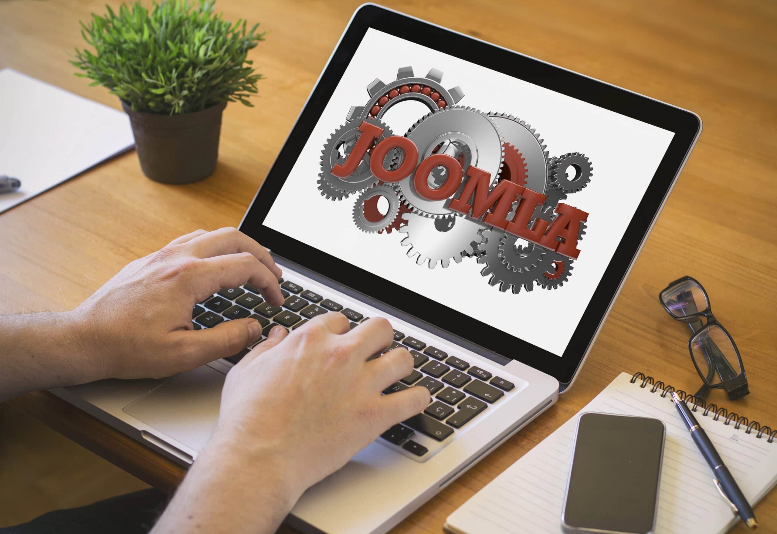 A person is typing on a laptop displaying the Joomla logo and gears, likely planning a content transfer to WordPress. A plant, notebook, and pen are neatly arranged on the wooden desk.