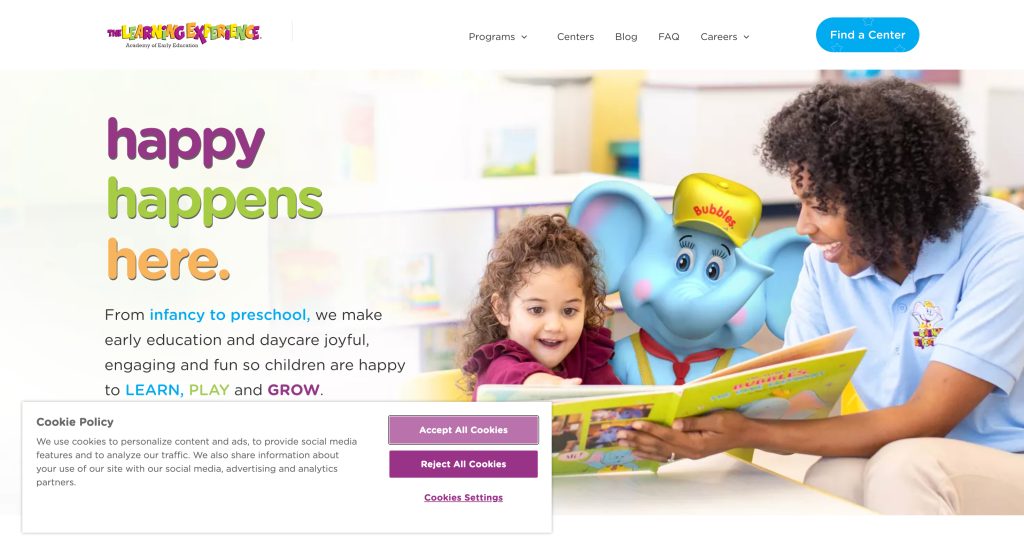 A smiling adult and young girl read a colorful book together beside a large blue elephant mascot in a bright daycare center, exemplifying the joyful spirit found on the best daycare websites. Text reads: happy happens here. Cookie policy pop-up is visible below.