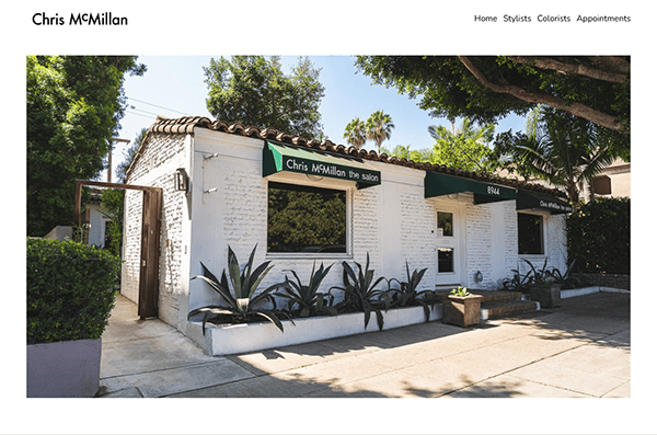 A white brick building with green awnings labeled "Chris MCMillan the salon" and "8944," surrounded by trees and plants, is where the best hair stylist in the area creates magic, bringing visions to life like those featured on top hair stylist websites.