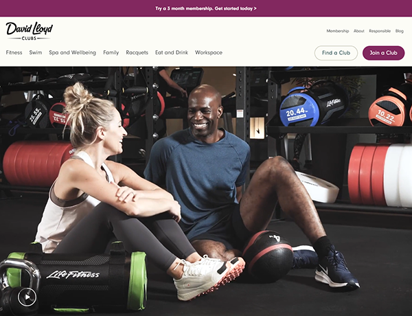 Two people are sitting on a gym floor, surrounded by fitness equipment, casually chatting and smiling. They exchange tips on top gym websites that inspire their workouts and help them stay motivated.