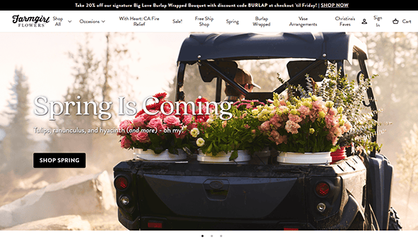 A truck loaded with colorful flower bouquets on its back drives down a rural road. The text reads, "Spring Is Coming," inviting you to explore Farmgirl Flowers' spring collection, showcased beautifully on one of the best florist websites around.