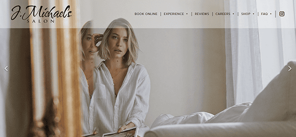 A woman with short blond hair gazes into a softly lit mirror, her white shirt complementing the serene ambiance. Text above reads "J. Michaels Salon," known for hosting the best hair stylist in town, with a sleek menu bar overlaying the reflection.
