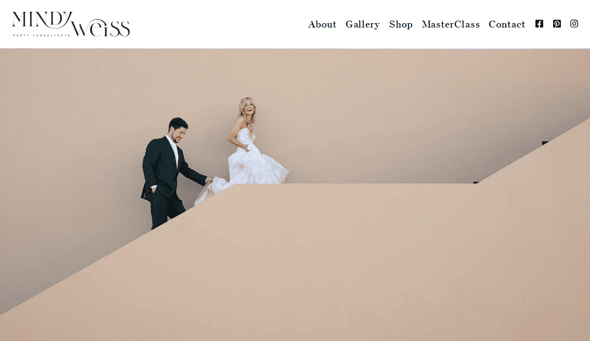 A bride in a white gown and groom in a dark suit walk up a beige, minimalist staircase against a plain wall, both smiling. This stylish, modern scene captures the elegant aesthetic seen on the best event planner websites.