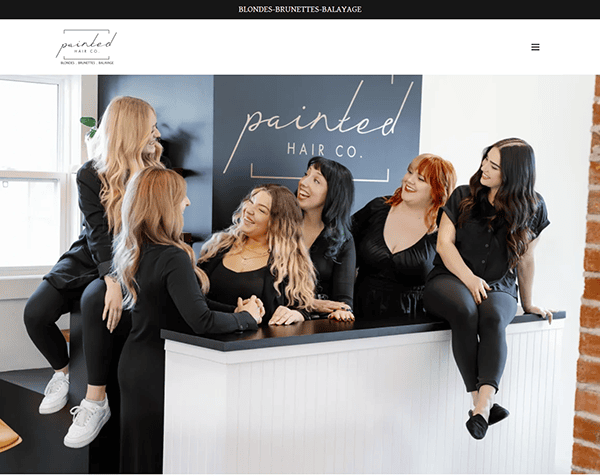 Six people in black outfits sit and stand around the salon reception desk with a "Painted Hair Co." sign, appearing to be smiling and chatting. It's clear they've mastered the art of styling hair, making them contenders for the title of Best Hair Stylist.