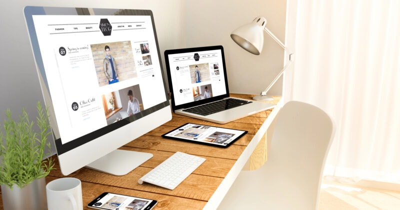 A modern wooden desk with a desktop computer, laptop, tablet, and smartphone displaying the same website, next to a potted plant and a desk lamp by a window with white curtains.