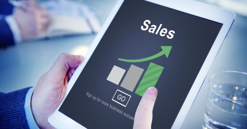 Person holding a tablet displaying a sales chart with a rising green arrow and bar graph. Another person's hand is visible, writing in a notebook. A glass of water is on the table.