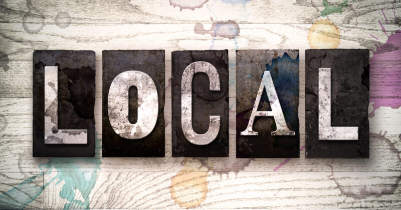 The word "LOCAL" is displayed in bold, weathered block letters against a wooden background with colorful paint splatters, evoking the vibrant spirit of local SEO and community-focused businesses.