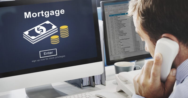 A man is on the phone at his desk, where a computer screen displays a mortgage application interface from one of the best mortgage websites. The interface includes dollar bills and coins icons, while another monitor shows a detailed spreadsheet.
