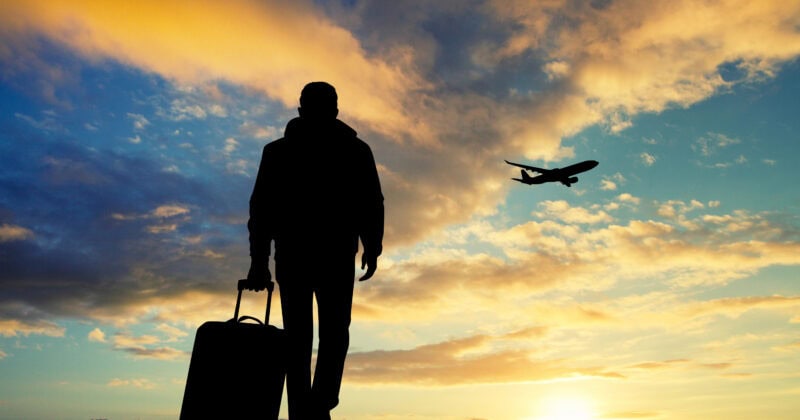 Silhouette of a person with a suitcase standing outdoors at sunset, watching an airplane fly in the sky.