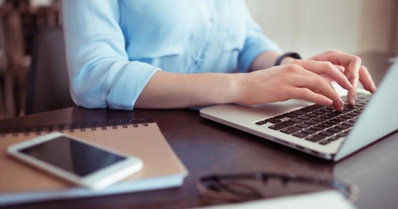 Person typing on a laptop at a desk with a notebook, smartphone, and eyeglasses nearby.