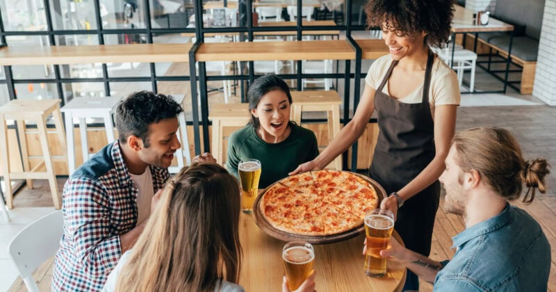 A waitress serves a large cheese pizza to four people sitting at a round table in a modern restaurant, each holding a glass of beer.