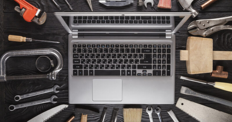 An open laptop, the focus of a project, sits surrounded by saws, wrenches, brushes, and clamps on a dark wooden surface—a scene reminiscent of laptop repair websites where digital solutions meet hands-on tools.