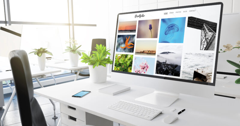 A bright office space with a large computer monitor displaying a photography portfolio. Nearby are plants, a smartphone, and office supplies on a white desk.