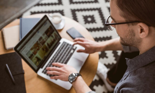 A person with glasses is using a laptop at a round wooden table with a notebook, smartphone, and coffee cup nearby on a patterned rug. The laptop screen displays a gallery of images related to WordPress and Squarespace domain setups.