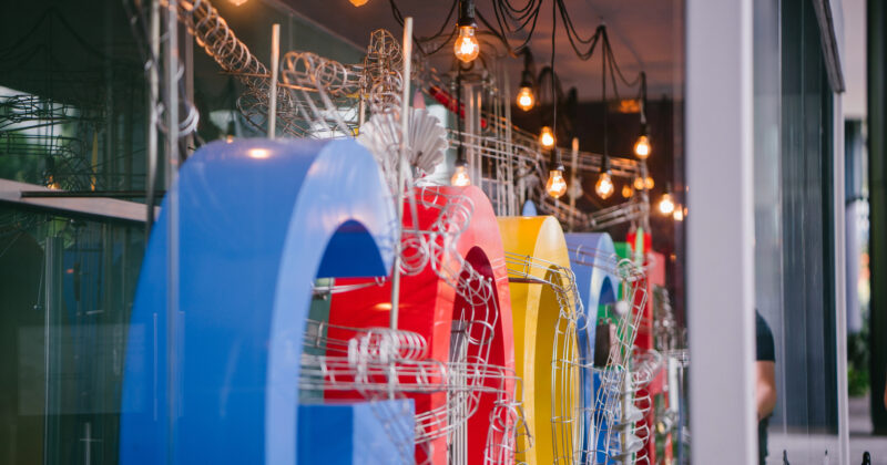 Colorful 3D letters spelling "Google" are displayed inside a glass-walled building, with hanging light bulbs above—an inspiring nod to search rankings and Google's Top 3 visibility.