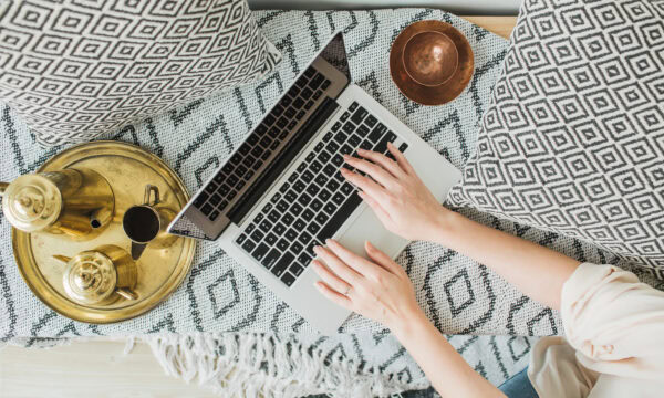A person types on a laptop, perhaps updating their WordPress site or planning to transfer a domain, while sitting on a patterned blanket with geometric pillows. By them, a brass tray with teapot and cups and a copper bowl create a cozy, relaxed atmosphere.