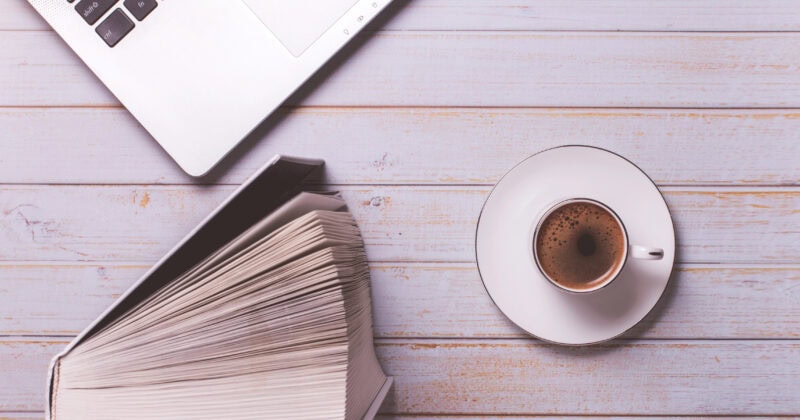 Open book and cup of coffee on a wooden surface, next to a laptop keyboard.