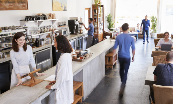 People in a cozy cafe, some engaged with laptops, perhaps working on social media strategies for small businesses. A barista assists a customer at the counter while another person walks by, and others are seated at tables in the background, enjoying the lively ambiance.