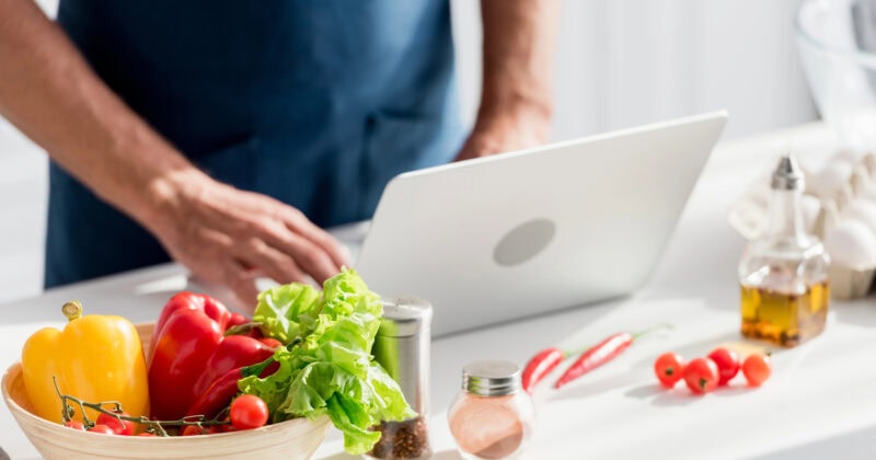 A person in a blue apron uses a laptop on a kitchen counter with vegetables, spices, and oil nearby.