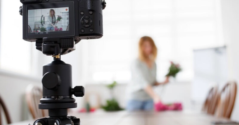 A camera on a tripod captures a person in the background, holding flowers inside a bright room, creating the perfect scene for a speed-optimized WordPress theme focused on blogging.