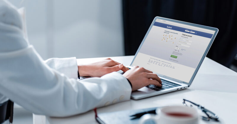 A person is using a laptop with a social media login page displayed, sipping coffee at a table. They're likely researching the best local service websites to connect with top-rated local service companies in their area.