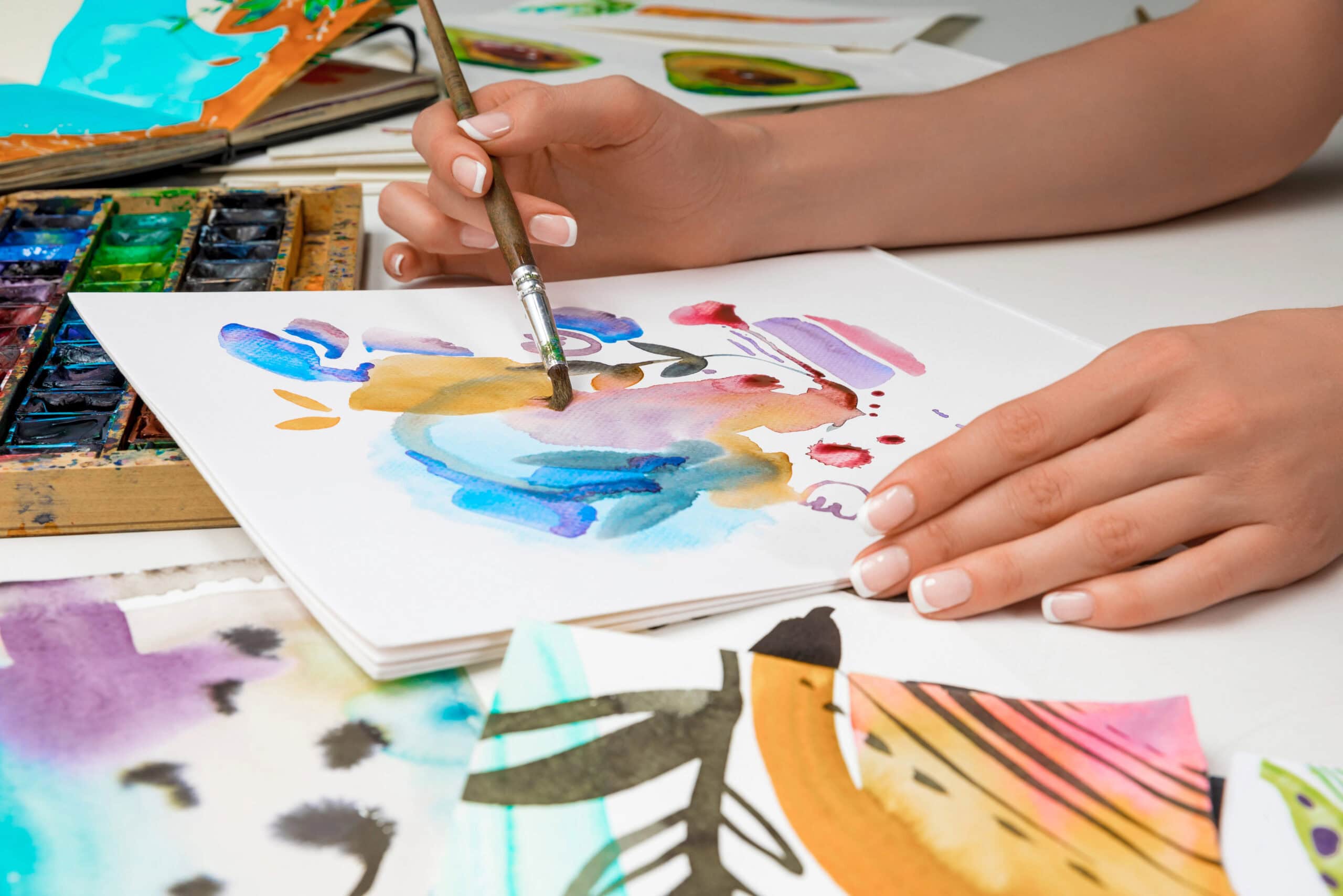 An artist skillfully paints an abstract watercolor design on paper, surrounded by a vibrant portfolio of colorful artworks and a lively palette.