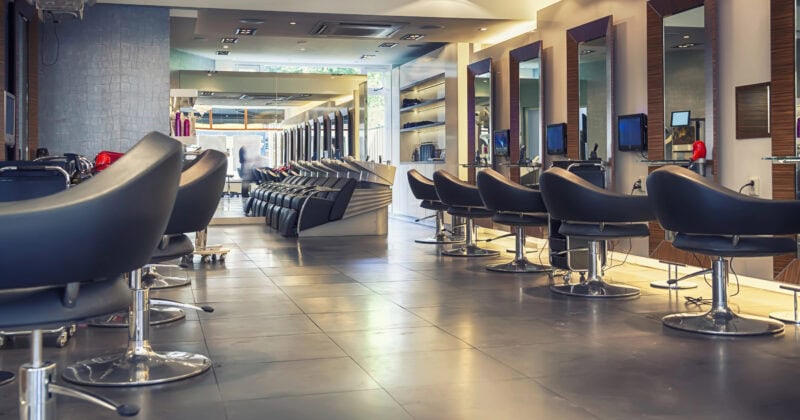 Modern, empty hair salon with black chairs, large mirrors, and washing stations, illuminated by ceiling lights and natural light from windows in the background.