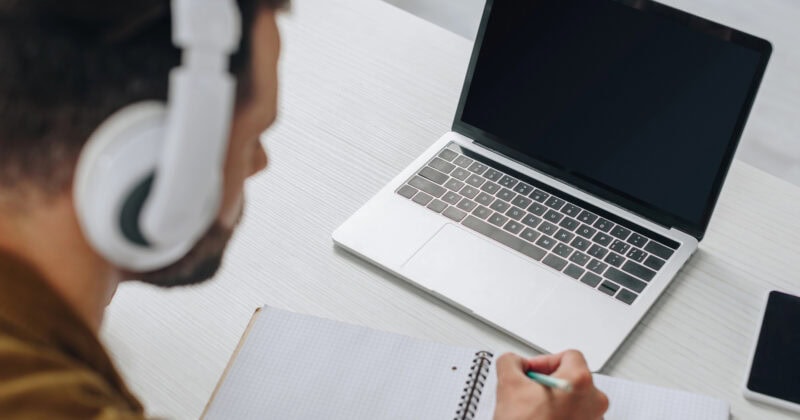 Person wearing headphones writes in a notebook while looking at an open laptop on a table.