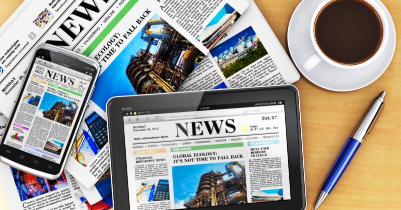 A collection of news-themed items on a table, including a smartphone, tablet, newspapers, a cup of coffee, a pen, and a pair of glasses.
