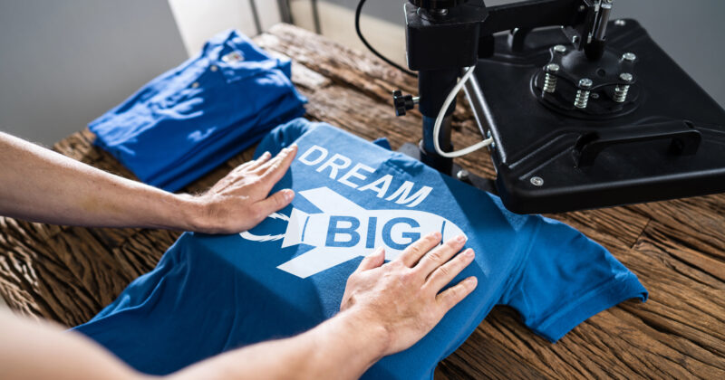 Person pressing a blue t-shirt with the text "DREAM BIG" using a heat press machine on a wooden table, showcasing one of the best t-shirt design creations; folded shirts are visible in the background.