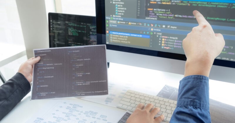 Two people are collaborating with computer code on a monitor and documents. One points at the screen while the other uses the keyboard, integrating user acquisition strategies. Various digital frameworks are visible as they blend software company insights into their work.