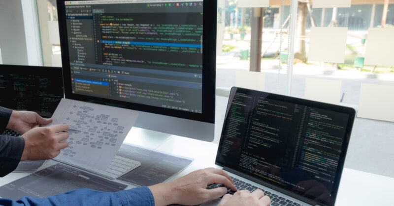 Two people work at a desk with a monitor and a laptop displaying code, while one holds a printed flowchart of a software design.