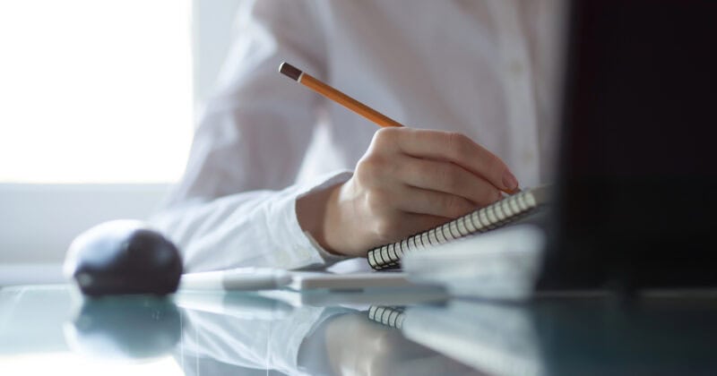 Person in a white shirt writing in a notebook with a pencil at a desk, next to a computer mouse and closed laptop—focused on an SEO audit to highlight SEO importance.