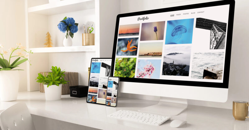 A home office desk hosts a large computer showcasing one of the best personal portfolios alongside a smartphone displaying stunning photography. The setup is complete with a potted plant, a camera, and organized shelves adorned with decor.