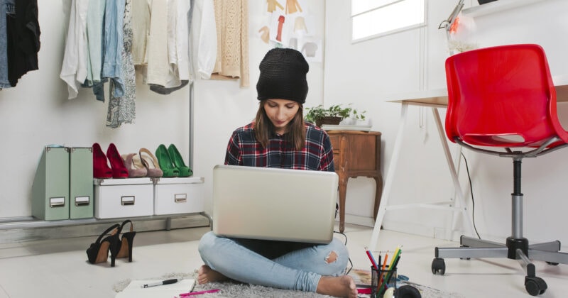 Woman sits on the floor in a modern room, working on a laptop surrounded by clothes, shoes, a desk, and scattered stationery.