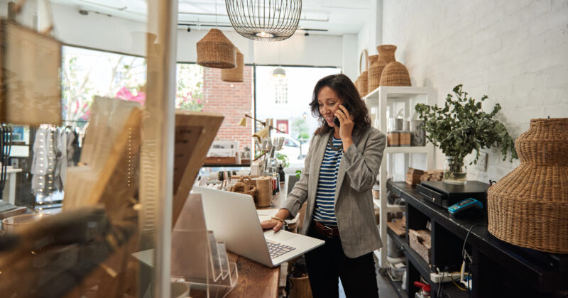 A woman stands in her boutique shop, talking on the phone and working on a laptop at the counter—balancing daily tasks as a small business owner. Woven baskets and decorative items from one of the best small business websites are displayed around her.