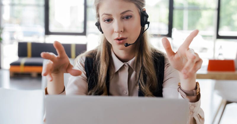 A woman wearing a headset gestures with her hands while sitting in front of a laptop, likely engaged in a video call.