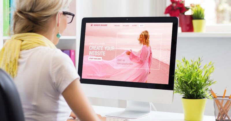 A person wearing glasses and a yellow scarf sits at a desk using a computer. The screen displays a website builder page with the text "Create Your Website" and a start building button.
